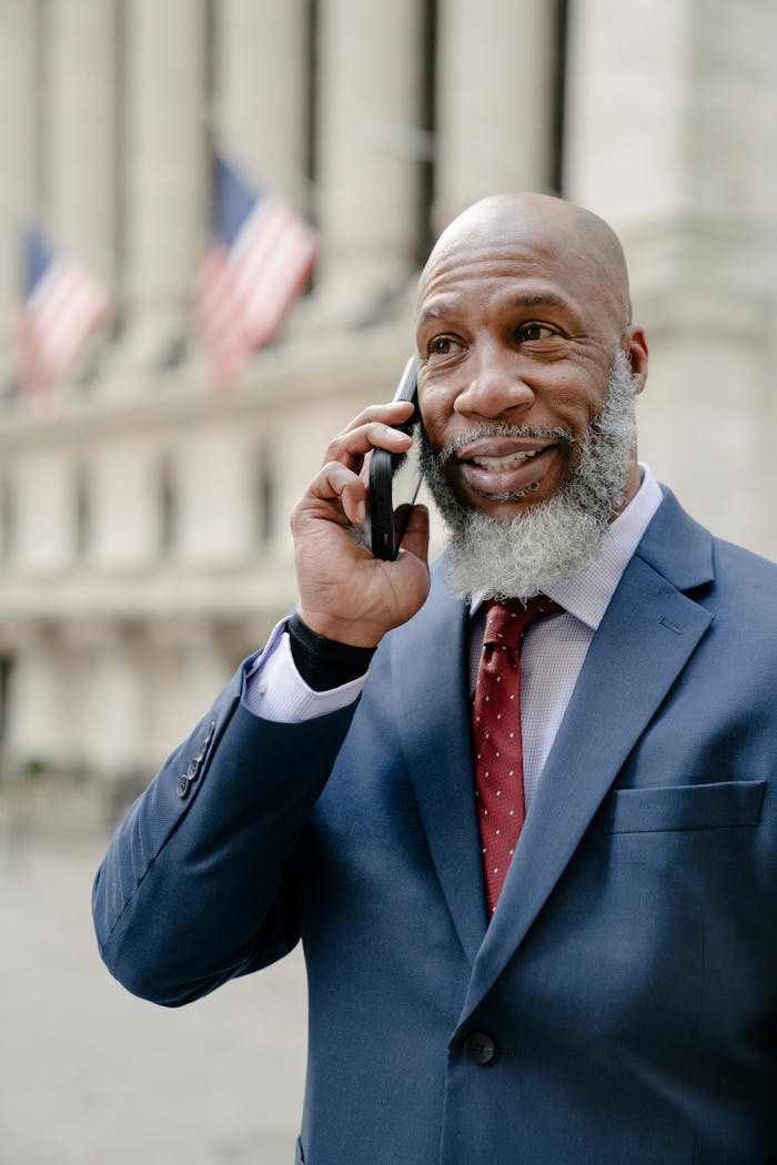 A professional man in a suit talks on his smartphone outdoors. Patriotic flags in the background.